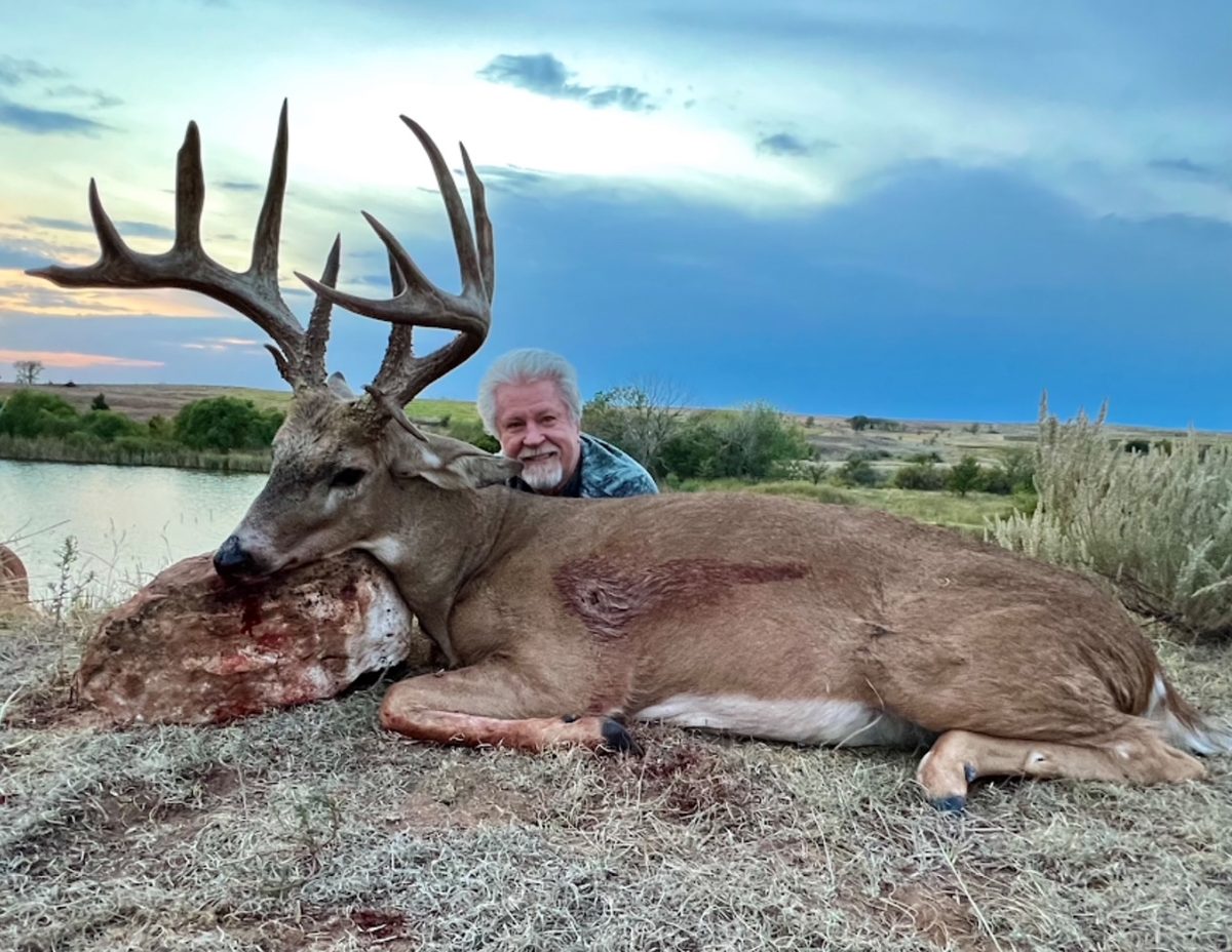 Hunter with a big-framed whitetail taken beside a stock pond at dusk